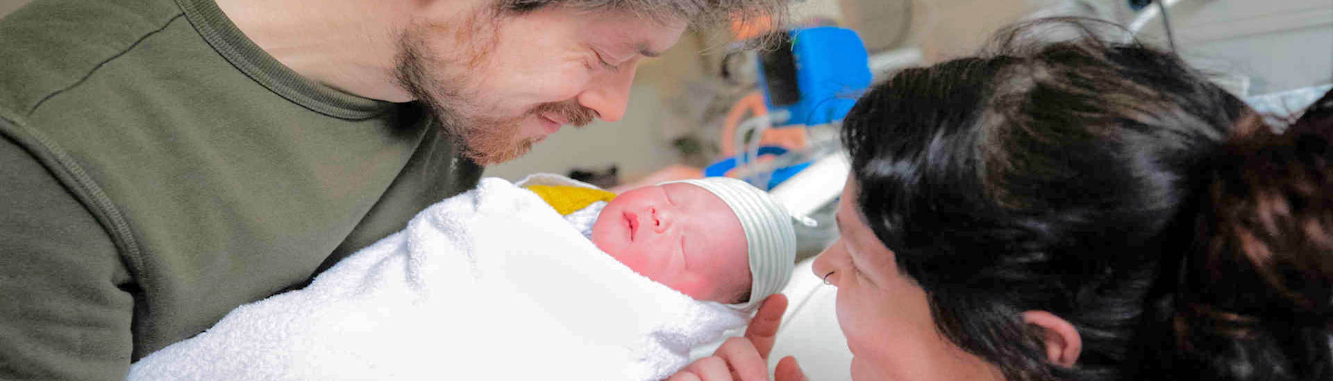 A smiling man and woman look at a newborn baby with medical equipment in the background